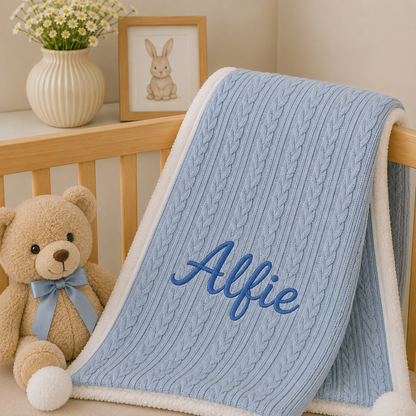 A personalized baby blanket draped over a wooden crib, featuring a light blue cable-knit pattern, a white sherpa border, and the name 'Alfie' embroidered in blue. A teddy bear and a vase of daisies are styled next to it.