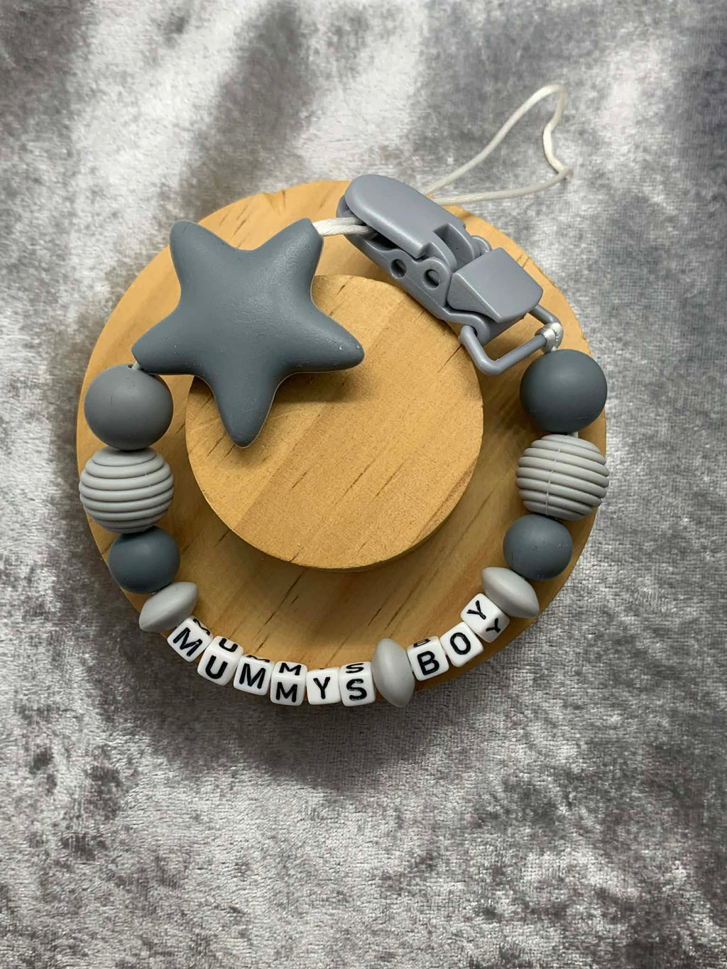 Dark Grey Silicone Dummy Clip with Dark Grey star personalised with the name 'Mummy's-Boy' on a grey textured surface. Can be personalised