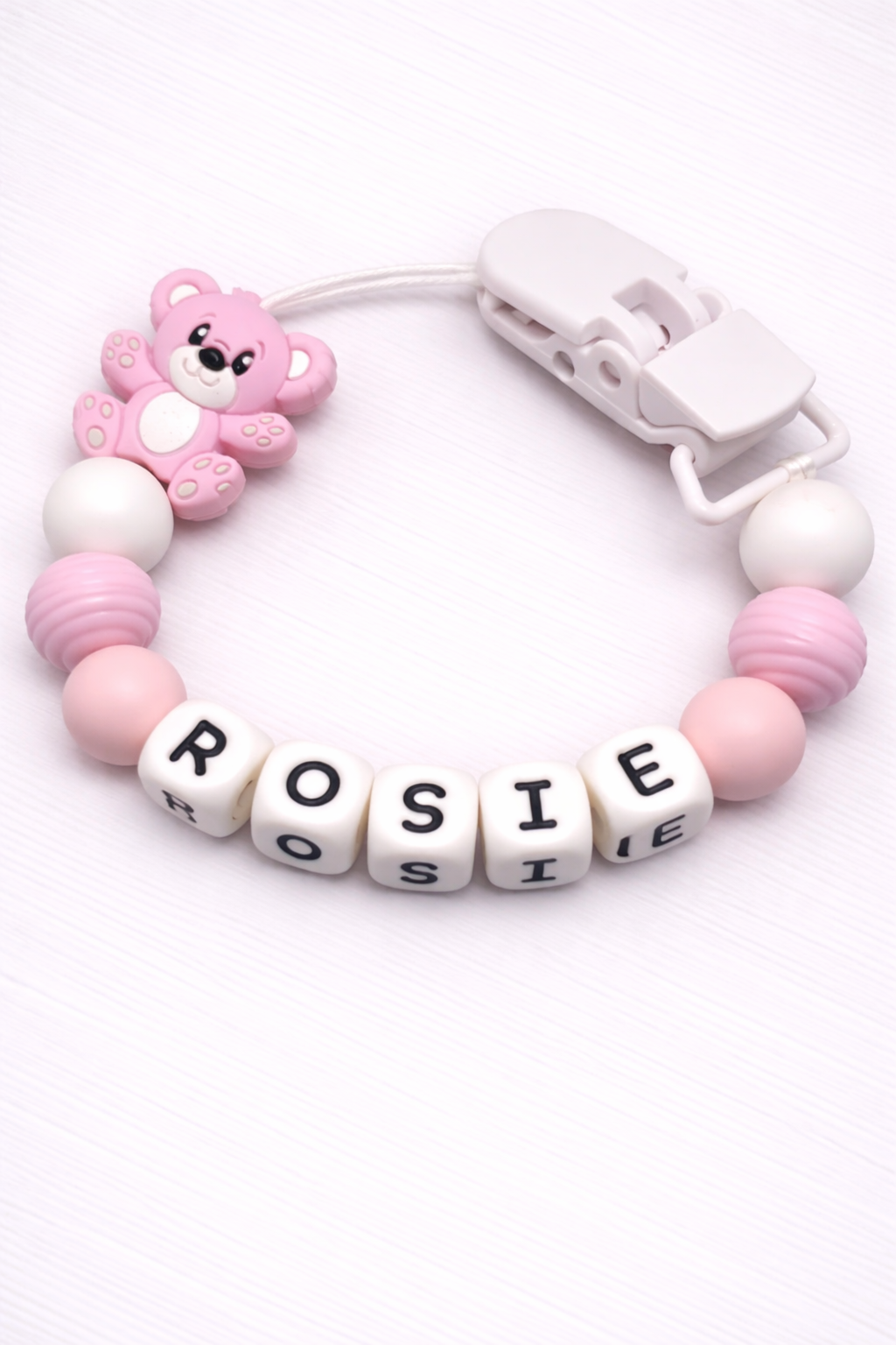 Pink and white baby teether with 'ROSIE' letters and a teddy bear charm on a white background