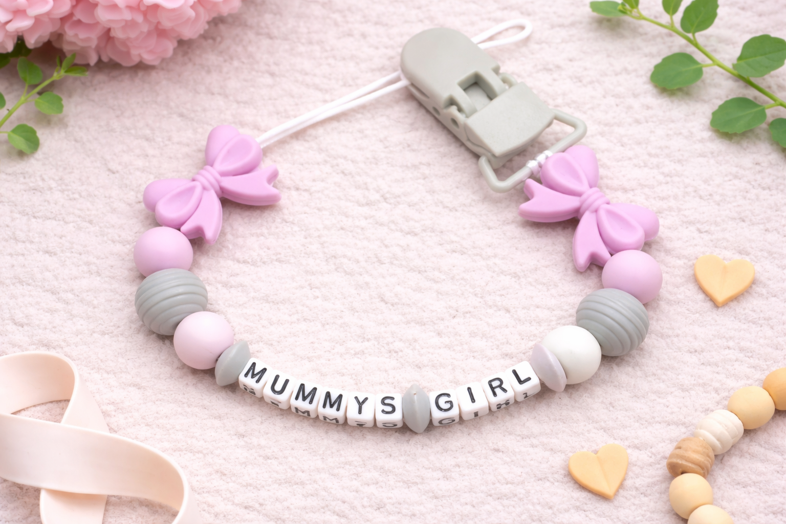 Flat lay of personalized baby pacifier clip with pink bows and pastel beads spelling “MUMMYS GIRL” on a soft blush textured background.