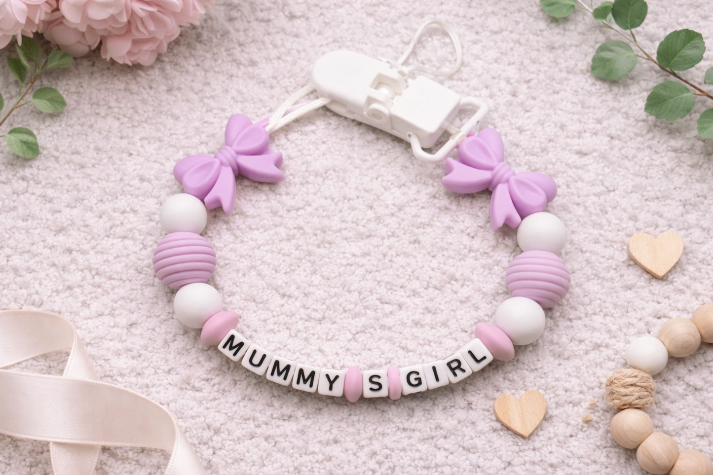 Personalized baby pacifier clip with lavender bows and silicone beads spelling Mummy’s Girl on a soft neutral textured background.