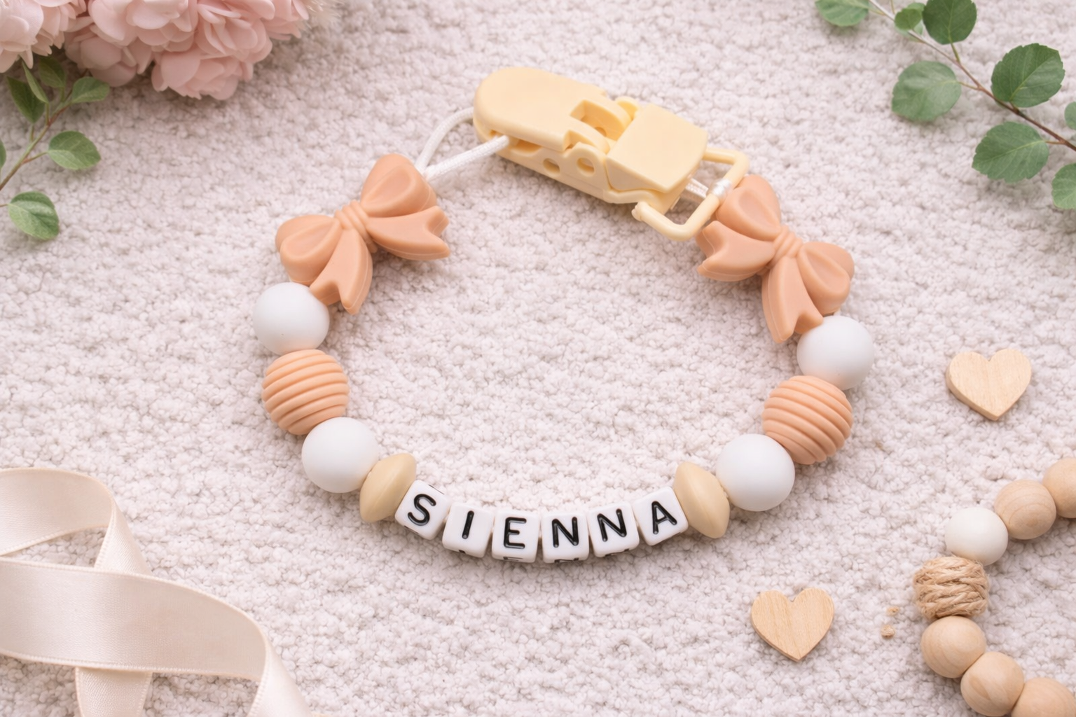 Personalized baby pacifier clip with peach bows and silicone beads spelling the name Sienna on a soft neutral textured background.