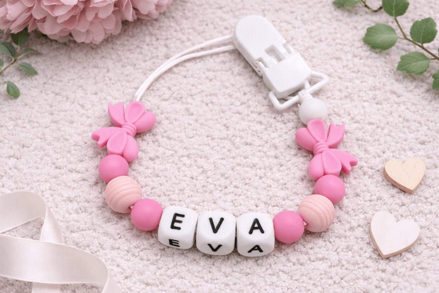 A beautifully styled flat lay of a personalized baby pacifier clip featuring soft pink silicone beads, bow accents, and white letter beads spelling “EVA.” Set against a cozy neutral fabric background with delicate floral and wooden heart details, this mockup highlights a sweet, feminine design perfect for baby gifts, announcements, or product listings.