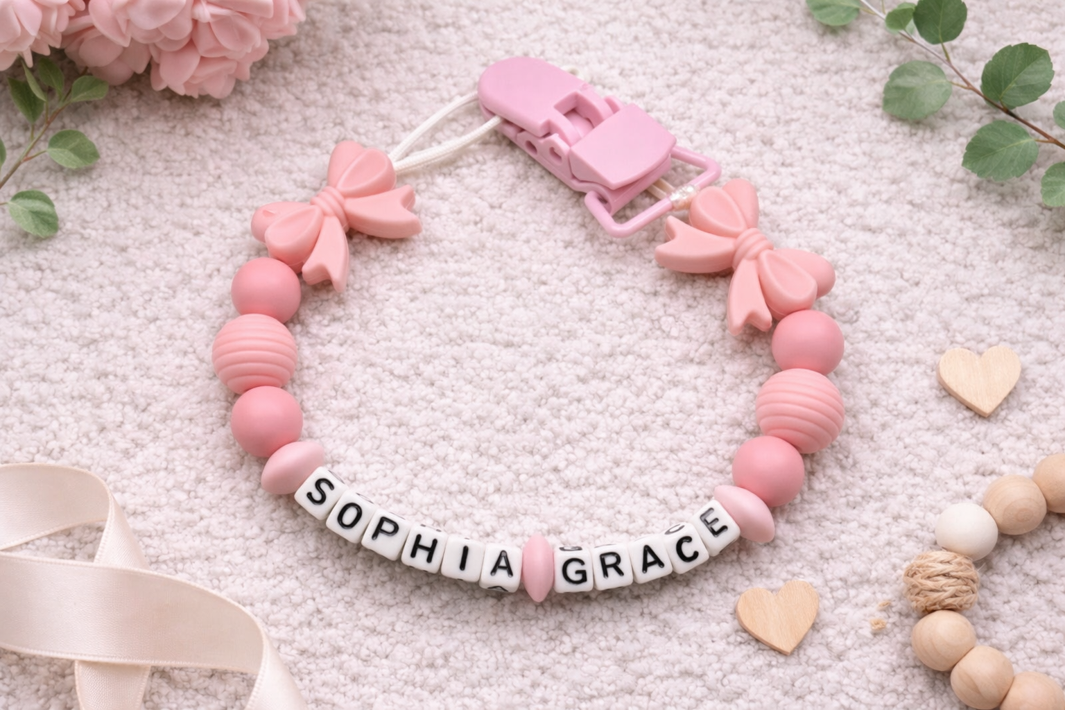 Personalized baby pacifier clip with pink silicone bows and beads spelling the name Sophia Grace on a soft neutral textured background.