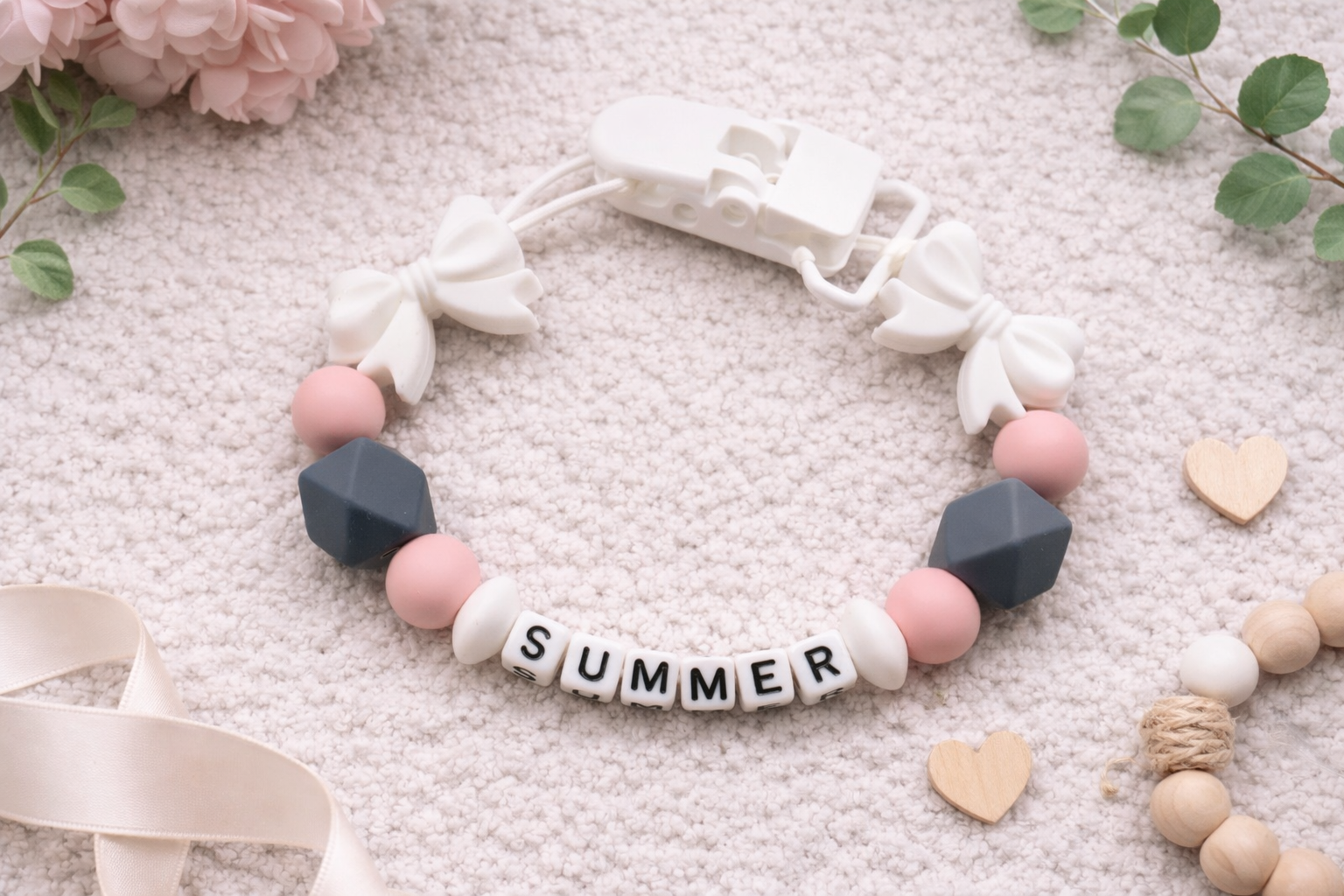 Personalized baby pacifier clip with white bow accents, pink and grey silicone beads, spelling the name Summer on a soft neutral textured background.
