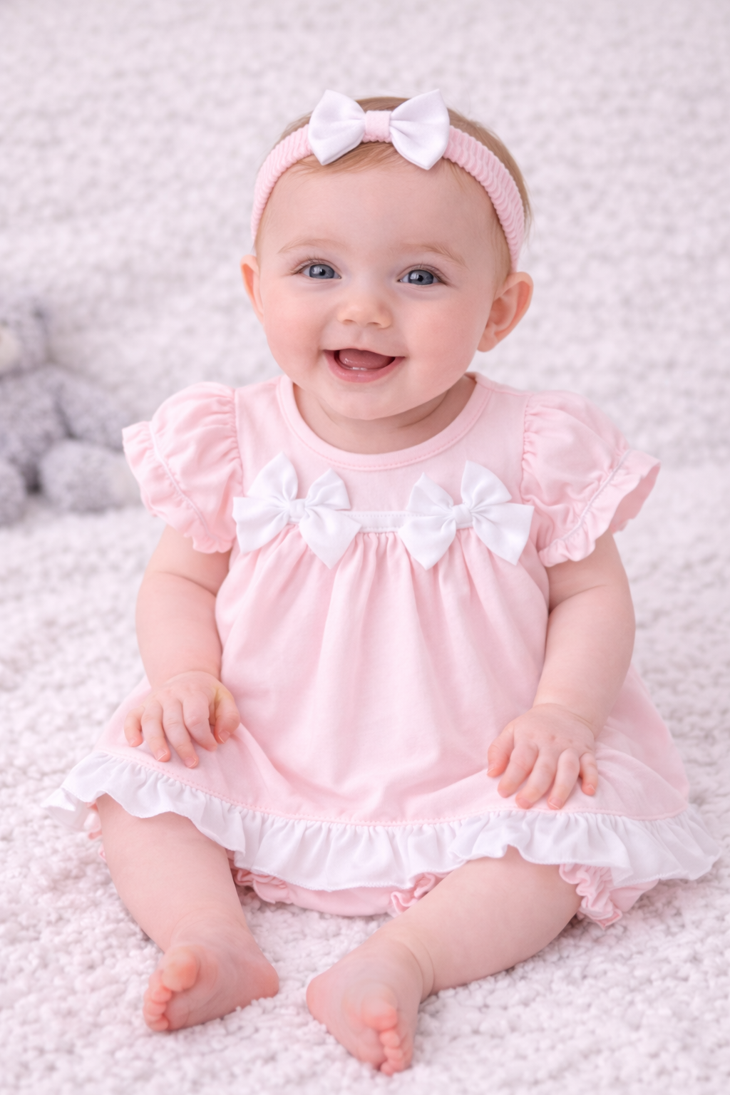 Baby in a pink dress with ruffles and a white bow headband on a soft surface.