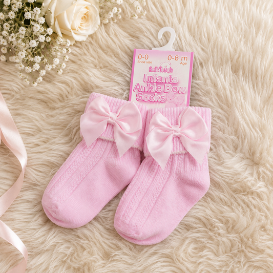 Pink baby socks with bows on a fluffy surface, accompanied by a packaging tag.