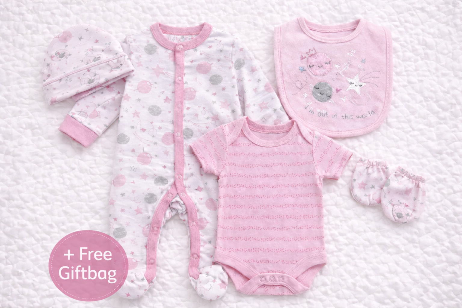 Flat lay image of a coordinated pink baby girls five-piece clothing set on a white quilted background, featuring a star and moon patterned sleepsuit, matching short-sleeve bodysuit, embroidered bib, baby hat, and scratch mittens, with a pink circular badge in the bottom left reading “+ Free Giftbag.”