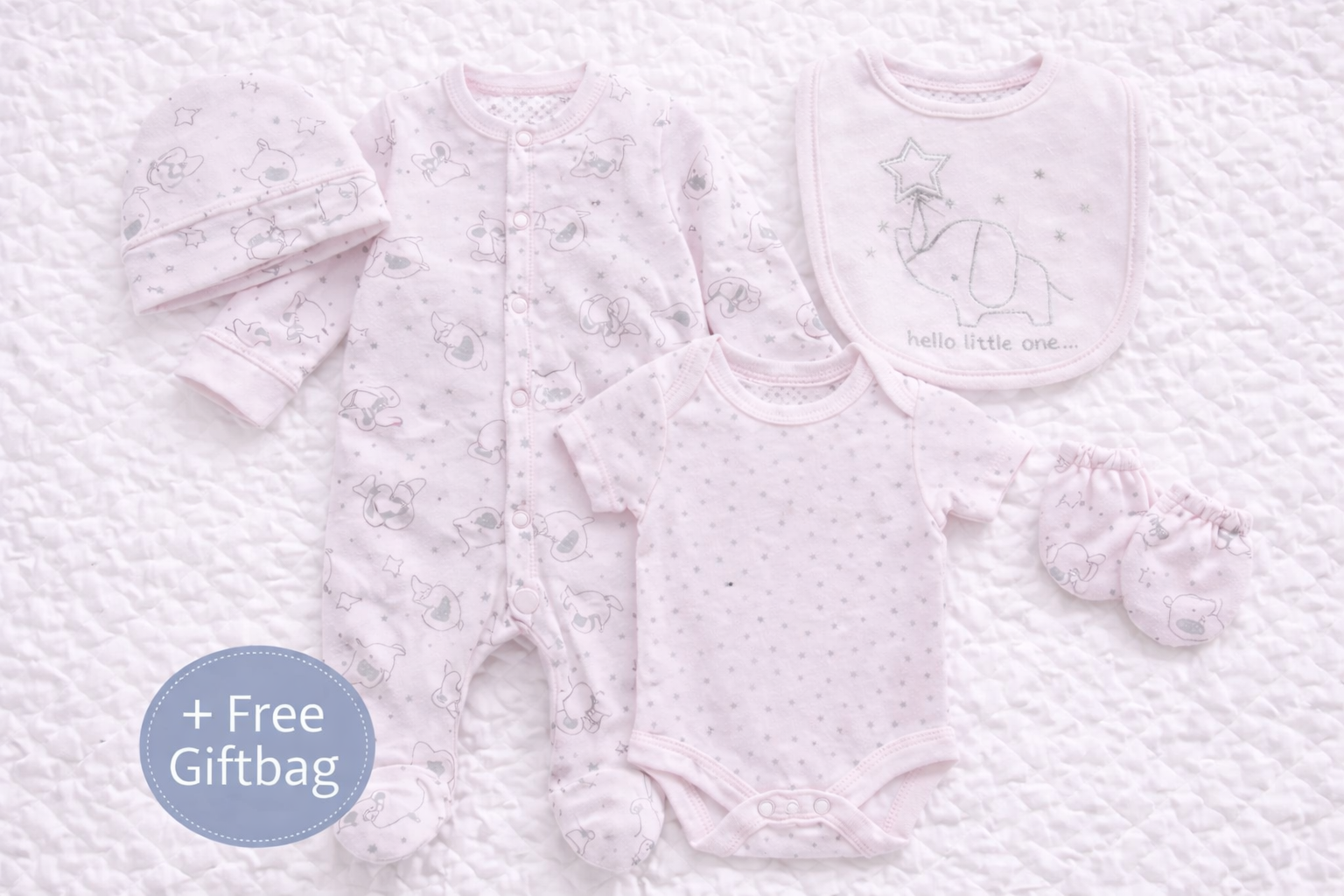Soft pink 5-piece newborn baby clothing set featuring an elephant and star print. Includes a footed babygrow, short-sleeve bodysuit, matching bib with embroidered elephant design, baby hat, and scratch mittens, all arranged on a white quilted background.
