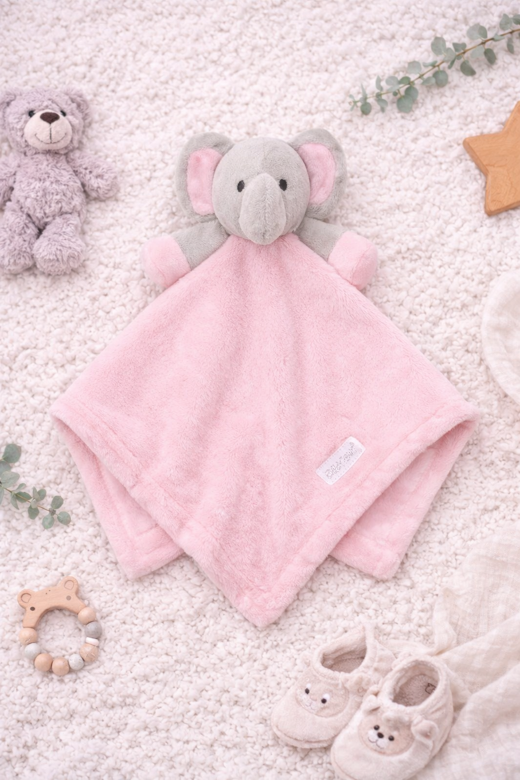 Soft pink elephant baby comforter displayed in a styled flat lay on a neutral cream nursery blanket, surrounded by baby accessories including booties, wooden toys, and greenery.