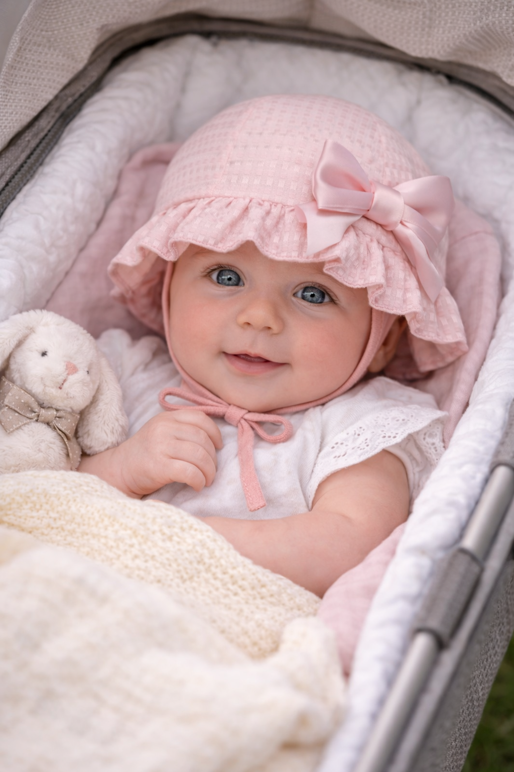 Baby in a pink bonnet sitting in a crib with a teddy bear.