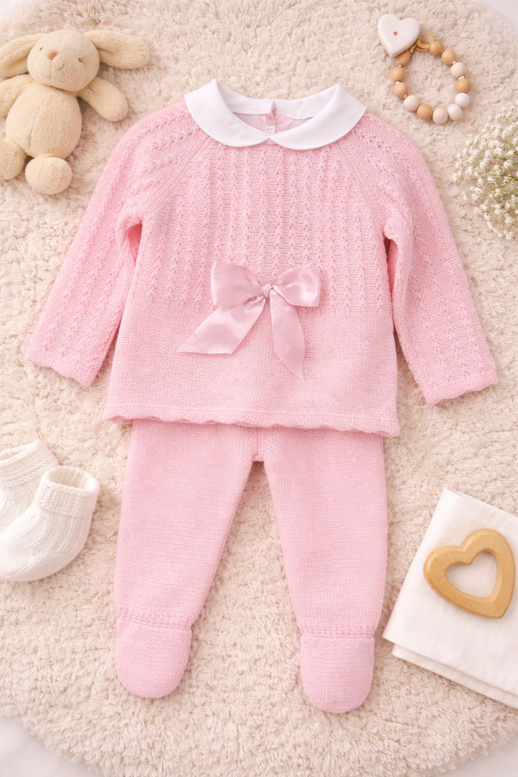 Infant dressed in a blush pink knit baby outfit featuring a classic collar, delicate bow detail, and matching trousers, perfect for newborn wear.