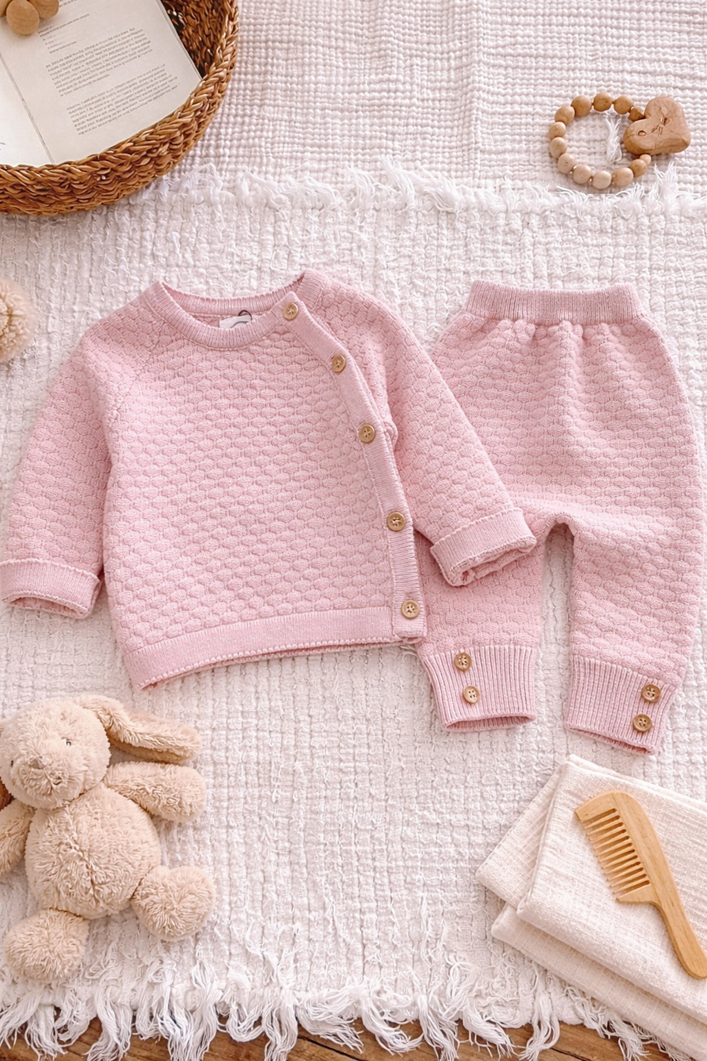 Newborn baby dressed in a blush pink knit two-piece outfit featuring a buttoned cardigan and trousers, photographed in a cosy neutral setting.