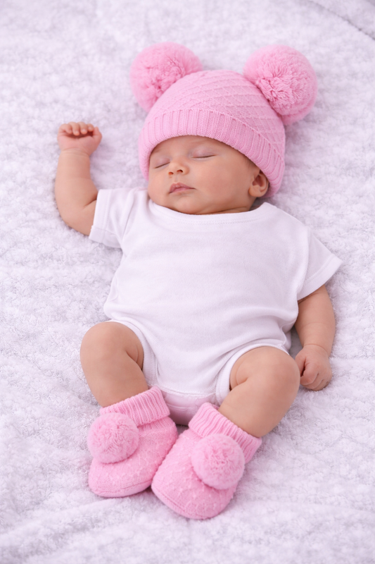 A sleeping newborn baby wearing a pink knit hat with two pom-poms, a white short-sleeve bodysuit, and matching pink booties with pom-poms, lying on a soft white blanket.