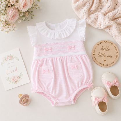 Pink baby romper with ruffled top and bow details on a light surface with flowers and cards.
