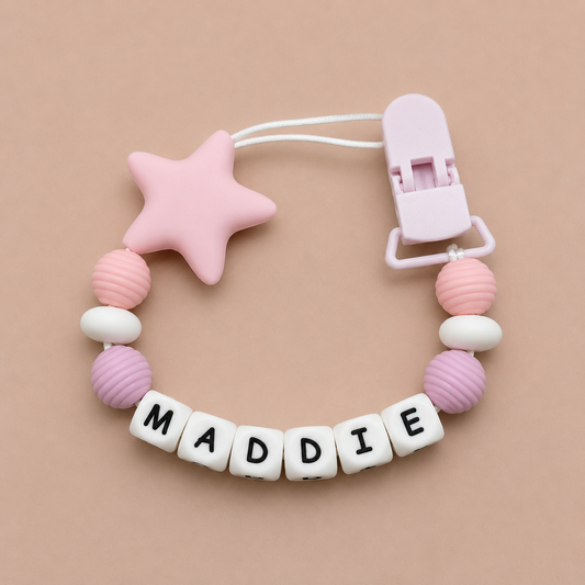 Pink and white pacifier clip with star-shaped bead and letter blocks spelling 'MADDIE' on a beige background