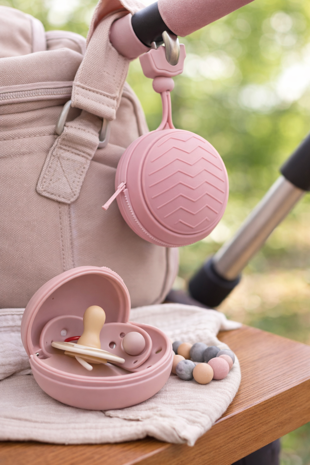 Cream silicone dummy holder clipped to a beige stroller bag, with a matching round pacifier case open on a wooden bench. The case contains a silicone pacifier and is styled with neutral-toned beads in a soft outdoor setting.