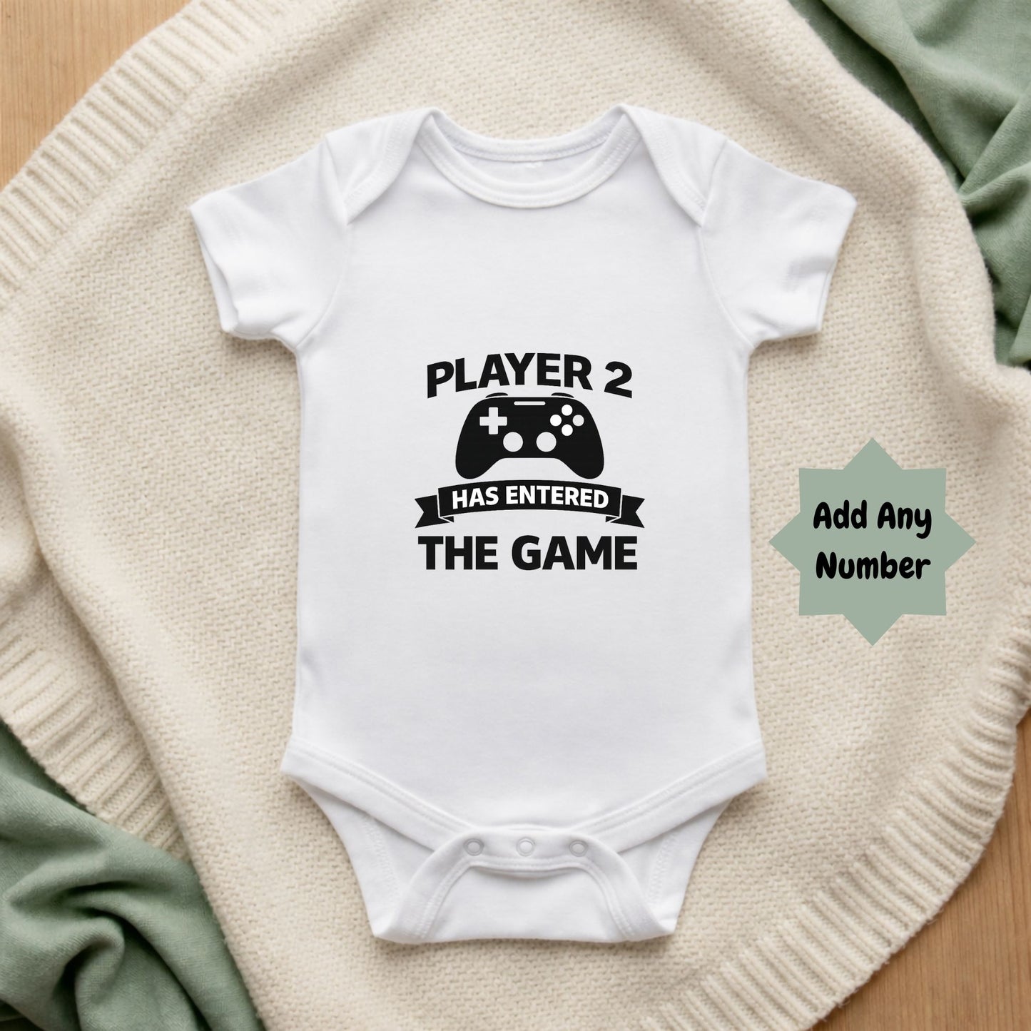 White baby onesie with 'Player 2 Has Entered The Game' text on a beige blanket.