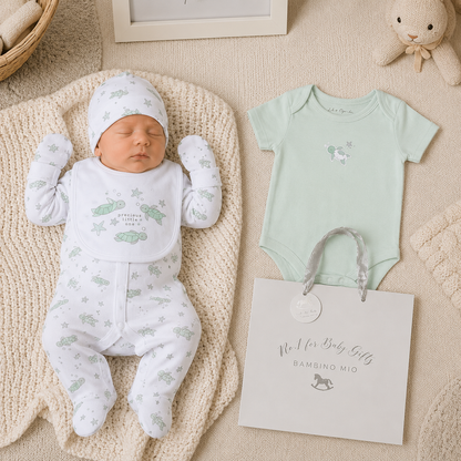Newborn baby in white outfit with green patterns, next to a light green onesie and gift bag on a textured surface.