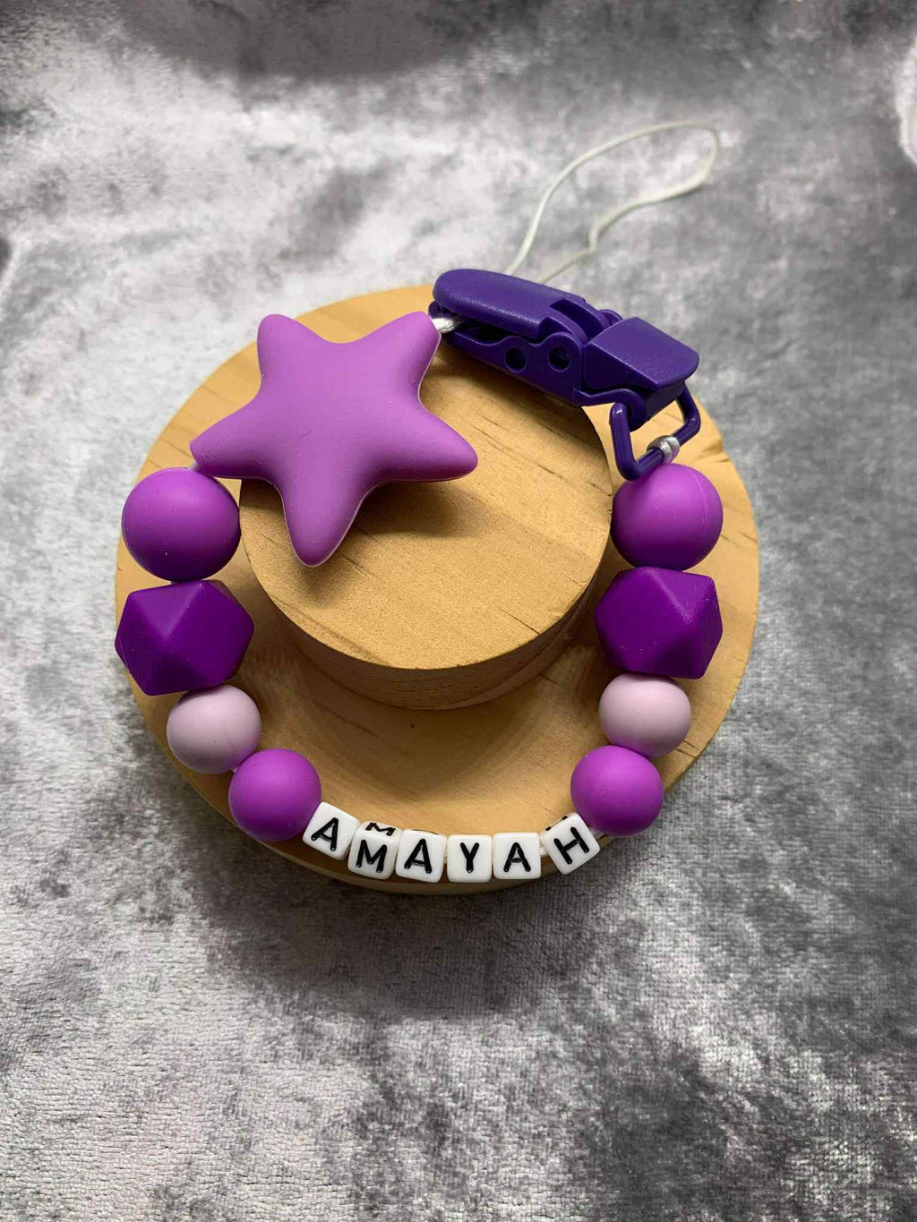 A beaded pacifier clip with purple beads, a purplw star, and letter beads spelling "Amayah" lying on a textured grey surface.