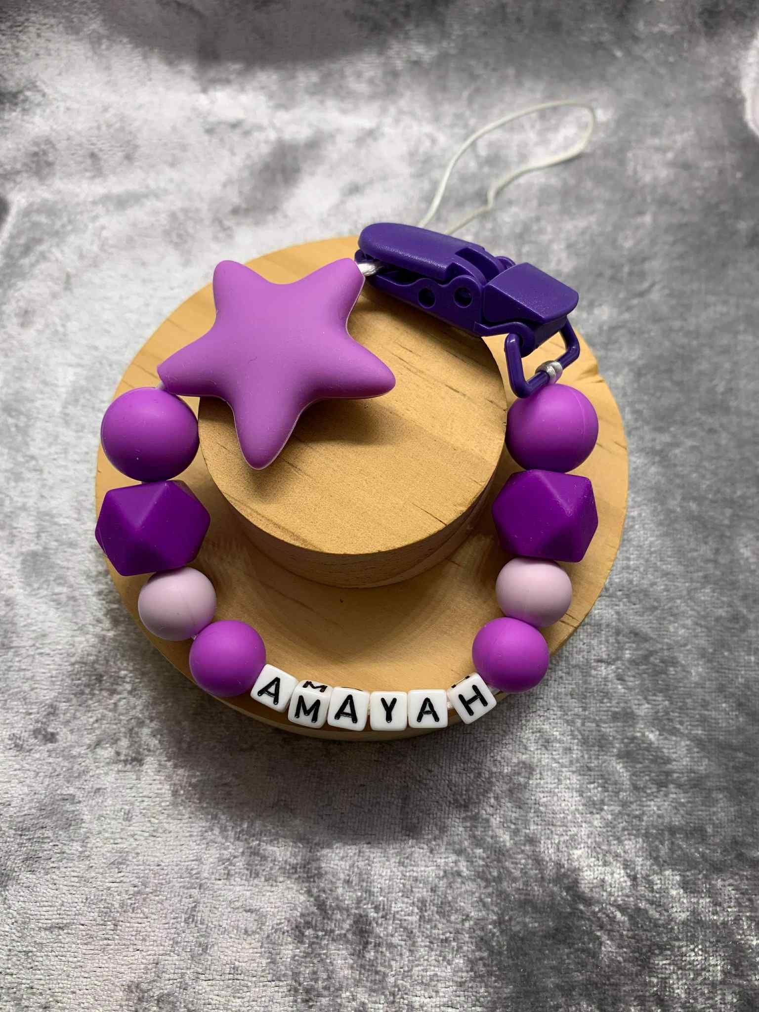 A beaded pacifier clip with purple beads, a purplw star, and letter beads spelling "Amayah" lying on a textured grey surface.