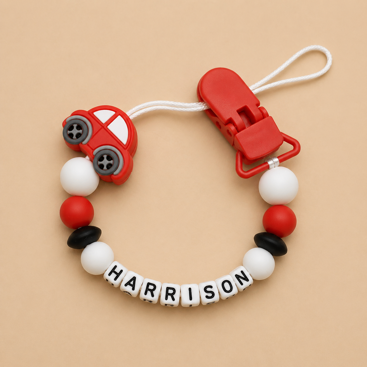 Baby dummy clip with red car design and 'HARRISON' name on a beige background