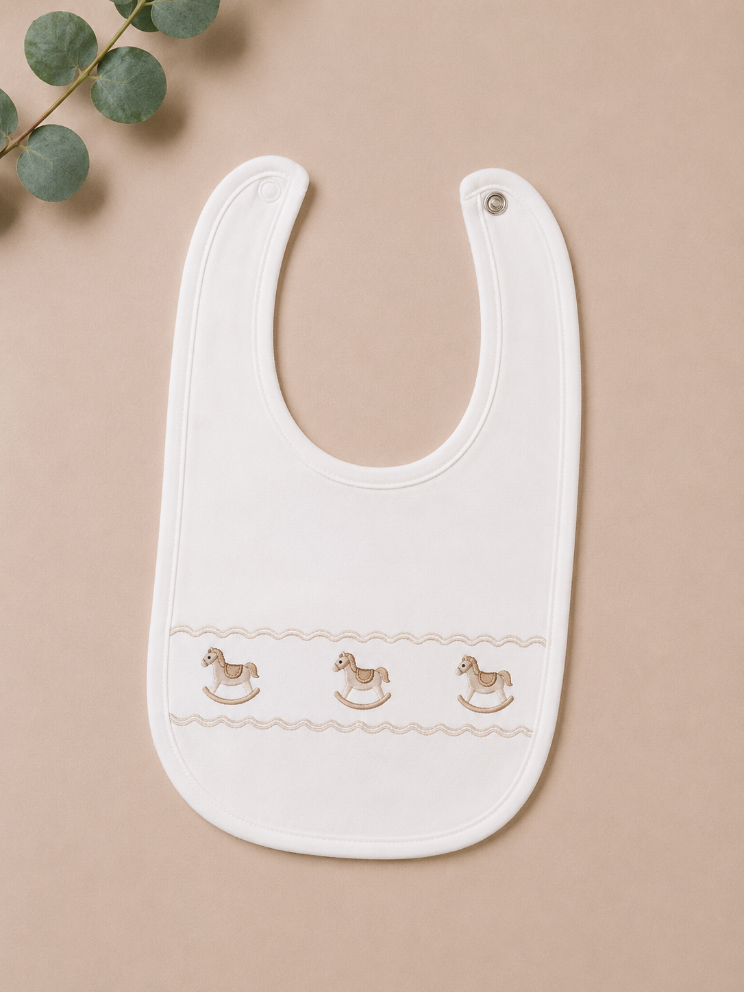 White baby bib with rocking horse embroidery on a beige background