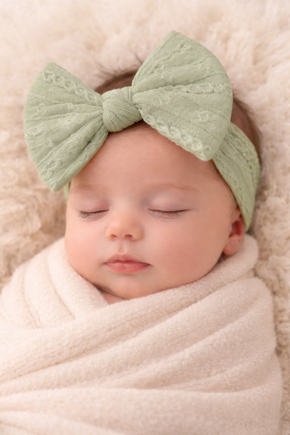 Newborn baby wrapped in beige blanket with green headband on light pink background