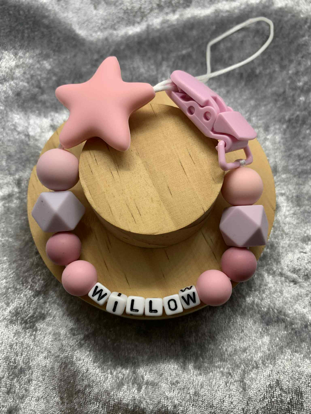A beaded pacifier clip with pink beads, a pink star, and letter beads spelling "Willow" lying on a textured grey surface. Personalised option available