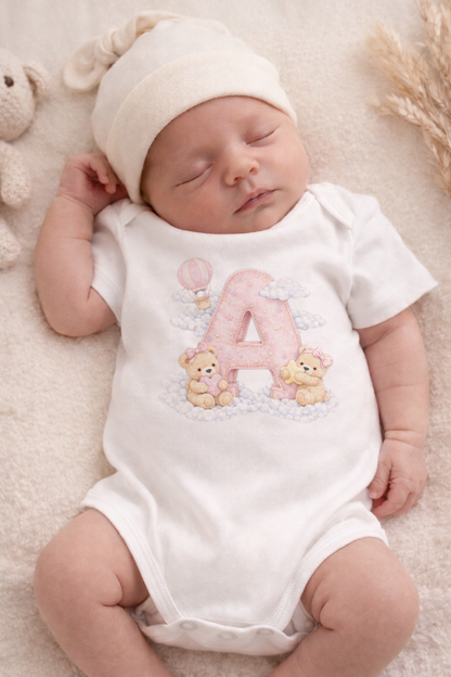 Newborn baby wearing a white onesie with a pink letter 'A' and teddy bears, lying on a soft surface.
