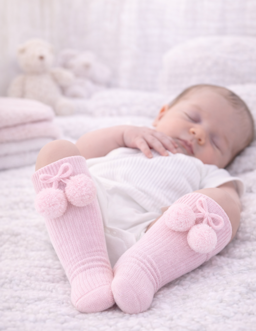 Baby wearing pink knitted socks with pom-poms on a soft surface