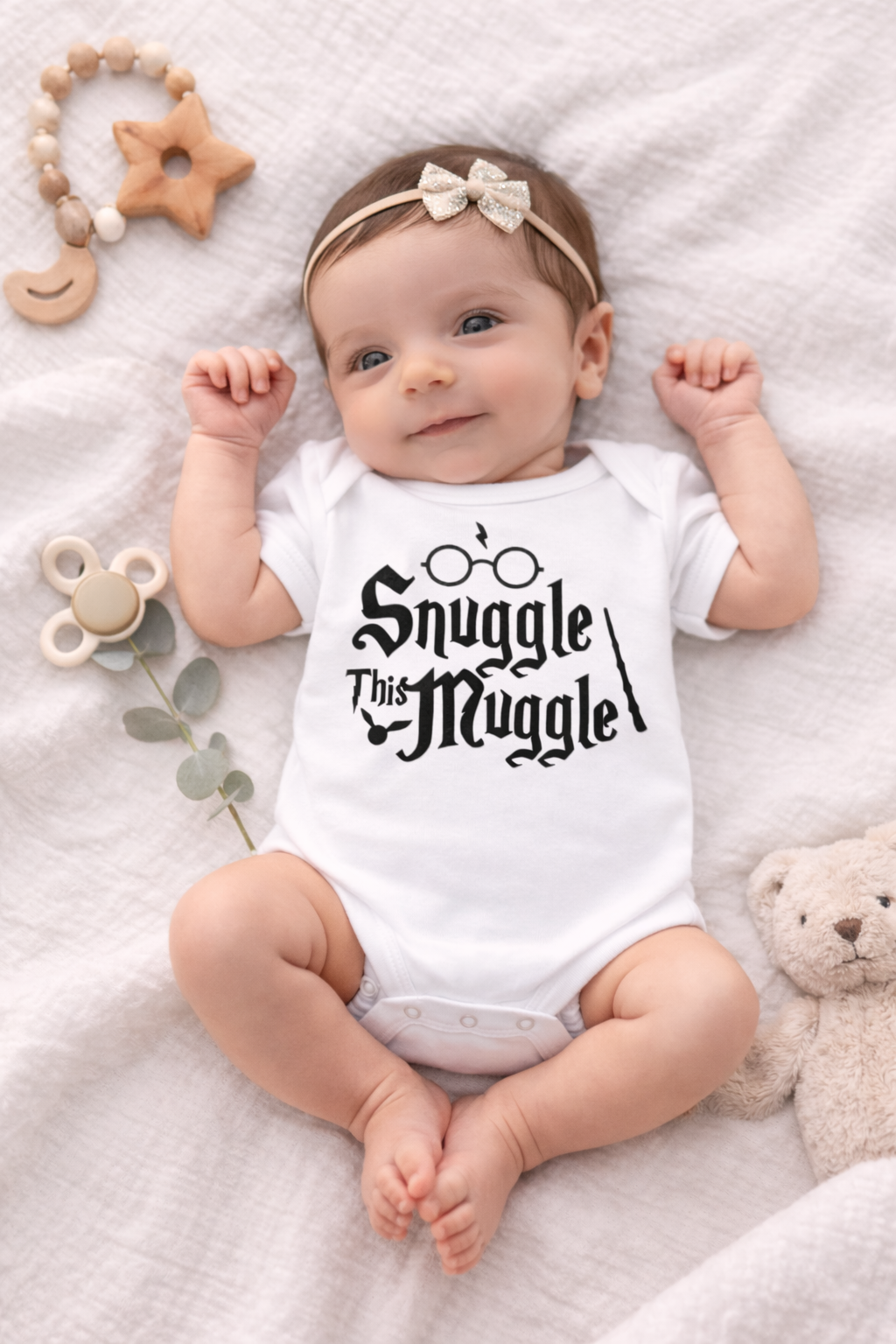 Baby wearing a onesie with 'Snuggle This Muggle' text, surrounded by toys on a soft surface.