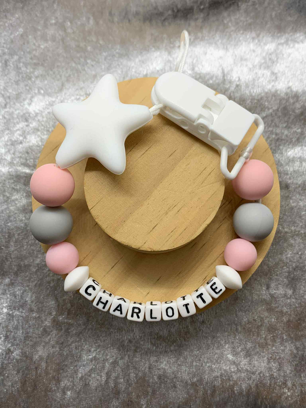 A beaded pacifier clip with pink, White and grey beads, a White star, and letter beads spelling "Charlotte" lying on a textured grey surface. Personalised option available