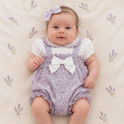 Baby wearing a purple floral romper with a white bow on a textured surface with lavender flower patterns.
