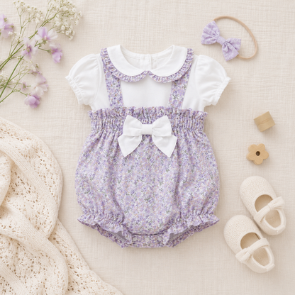 Lavender floral baby romper with white collar and bow, surrounded by baby items on a beige background.