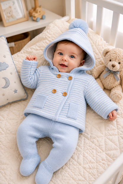 Baby in a blue knitted outfit lying on a crib with teddy bear and pillows.