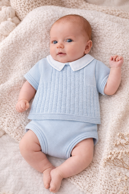 Baby wearing a light blue outfit with a white collar on a soft beige blanket.