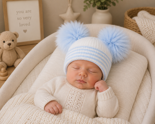 A sleeping newborn baby wearing a cream knit sweater and a light blue and white striped beanie with two large fluffy pom-poms, resting in a soft cream bassinet or chair. The background is a warm, cozy nursery setting with a teddy bear, framed quote, and small plant.
