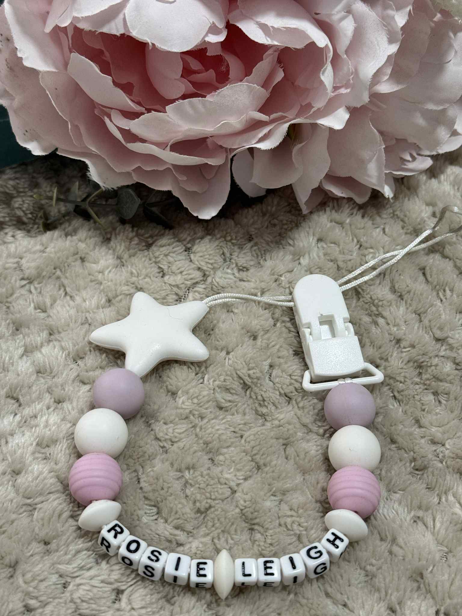 A beaded pacifier clip with pink, White and Lilac beads, a White star, and letter beads spelling "Rosie-Leigh" lying on a textured Beige surface. Personalised option available