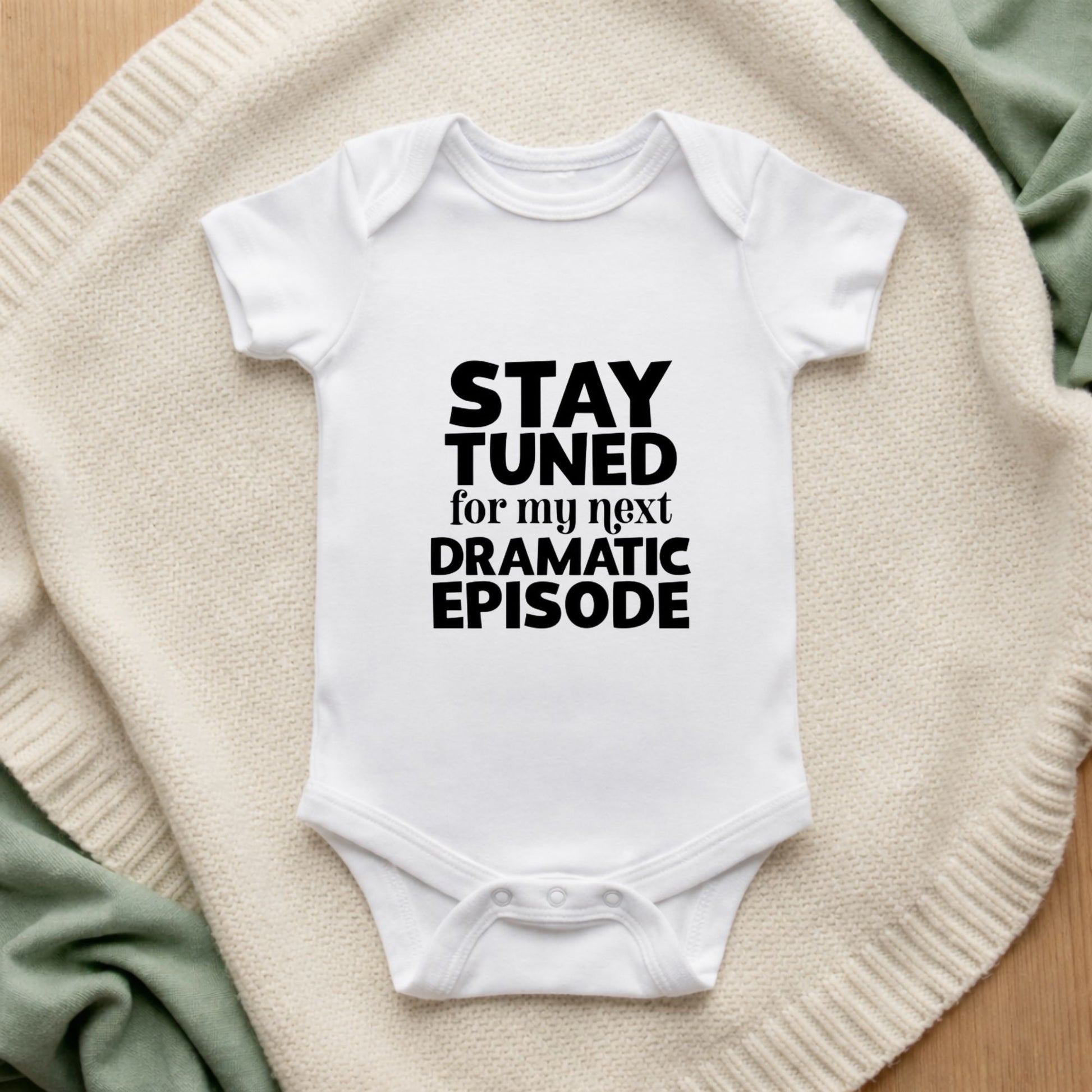 White baby onesie with text 'Stay tuned for my next dramatic episode' on a textured surface.