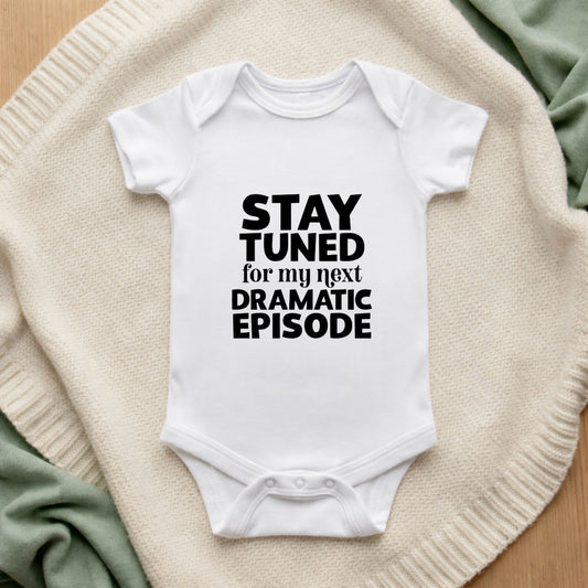 White baby onesie with text 'Stay tuned for my next dramatic episode' on a textured surface.