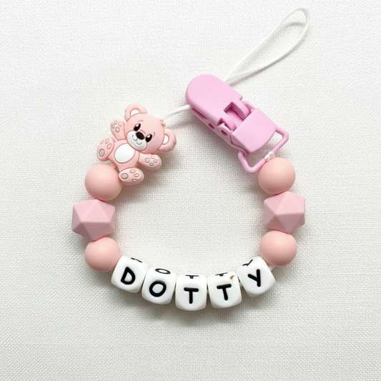 Personalised pink and peach teddy bear dummy clip on a soft white luxury linen textured background.