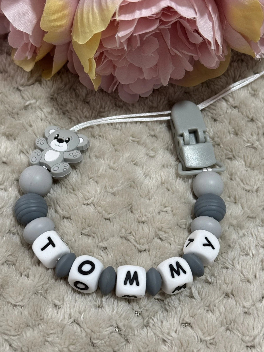 Dummy Clip with teddy bear charm and 'Tommy' beads on a textured surface with flowers in the background