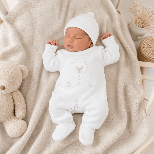 A sleeping newborn baby wearing a white long-sleeve footed onesie with a teddy bear face design and a matching white knit hat, lying on a beige blanket with a teddy bear and decorative props nearby.