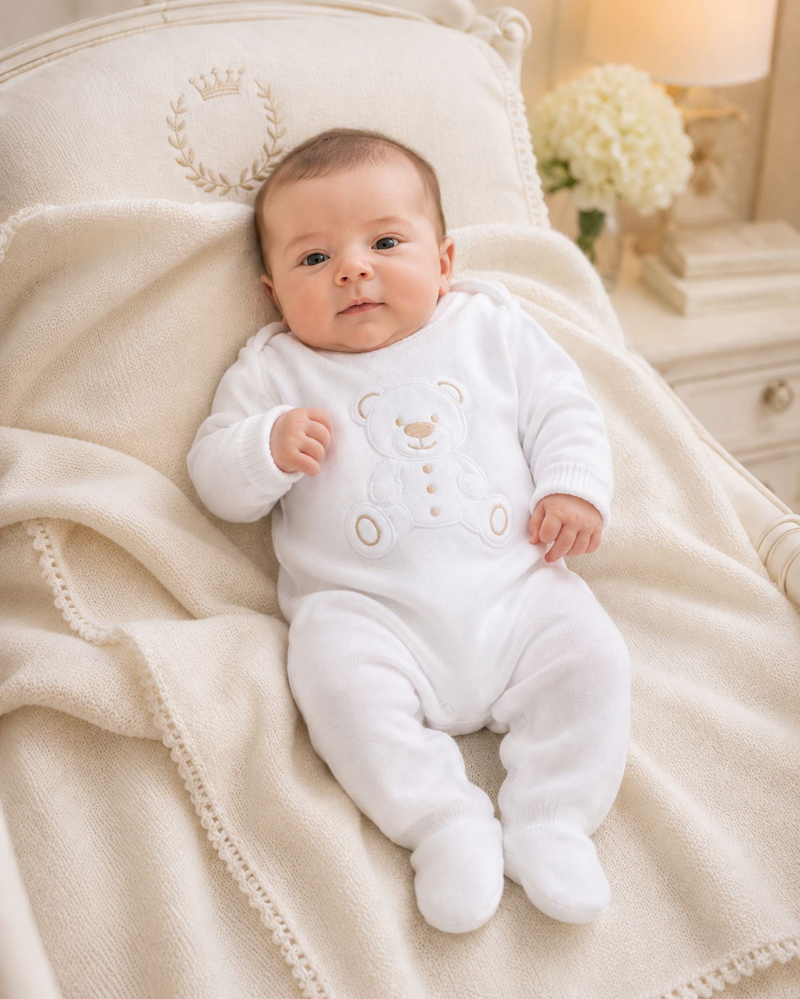 Baby in a white onesie lying on a soft surface with a decorative headboard in the background.