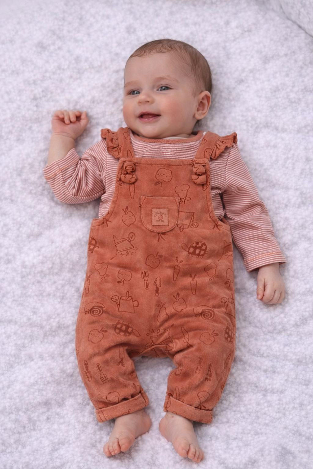 Baby wearing a pink long-sleeve shirt and brown corduroy overalls on a white textured surface