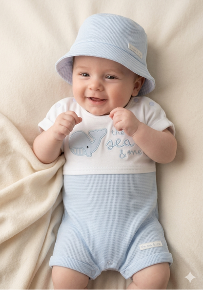 Baby wearing a light blue outfit with a matching hat, lying on a beige blanket.