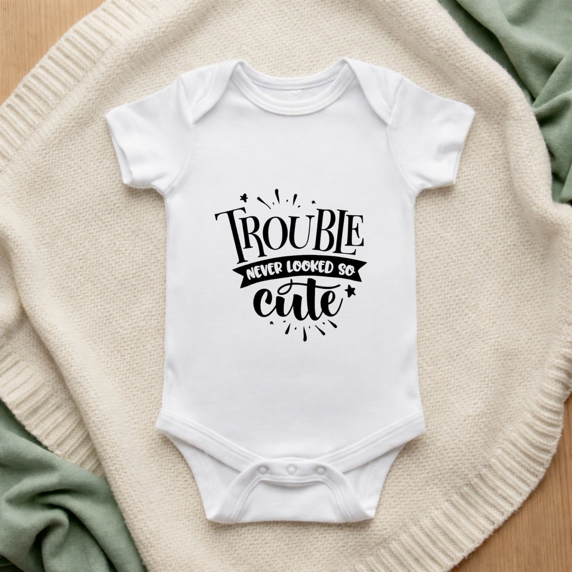 White baby onesie with 'Trouble never looked so cute' text on a textured surface.