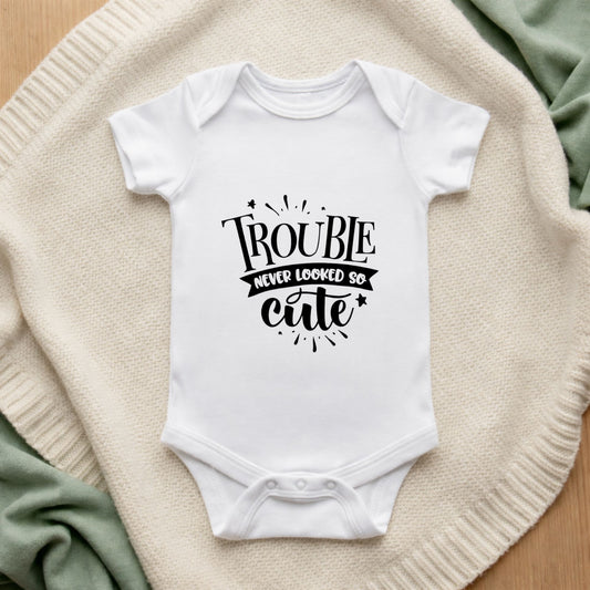 White baby onesie with 'Trouble never looked so cute' text on a textured surface.