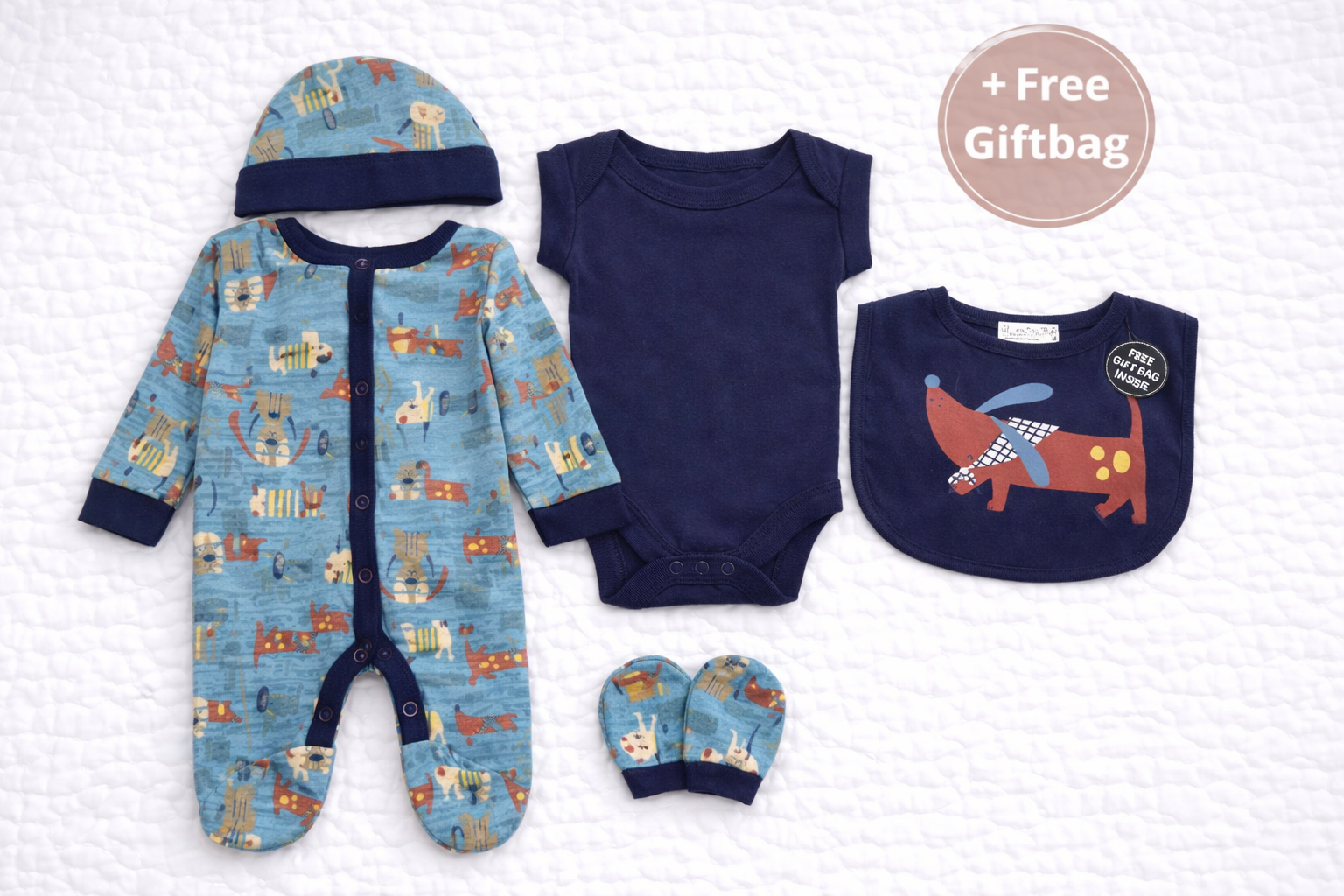 Flat lay of a unisex 5-piece newborn baby outfit set featuring a blue dog print sleepsuit, navy bodysuit, matching hat, coordinating bib with dog illustration, and mittens. The set is arranged on a soft white quilted background and includes a visible “+ Free Giftbag” circle, making it ideal as a newborn or baby shower gift.